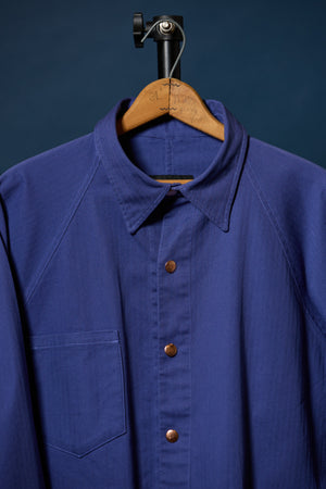 Barista Chore Coat in Washed Herringbonet on a wooden hanger against a dark background