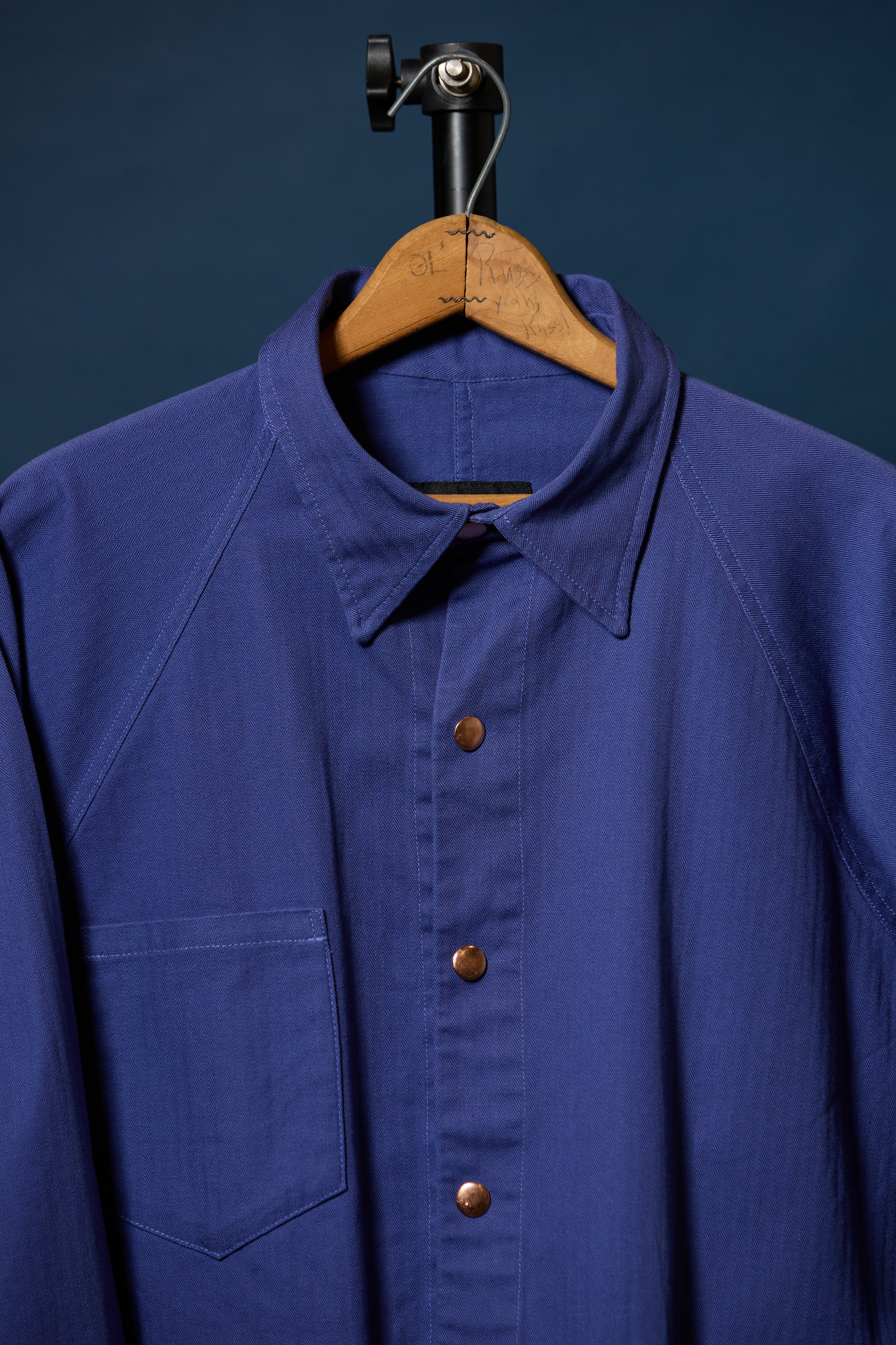 Barista Chore Coat in Washed Herringbonet on a wooden hanger against a dark background