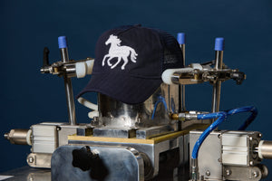 A navy blue trucker cap with a white embroidered horse logo is being held by a metal and pneumatic pressing machine against a dark blue background.