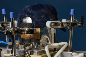 A navy blue mesh baseball cap is mounted on a transparent cylindrical mold inside a metal industrial press machine with blue-capped adjustment knobs, set against a dark blue background.