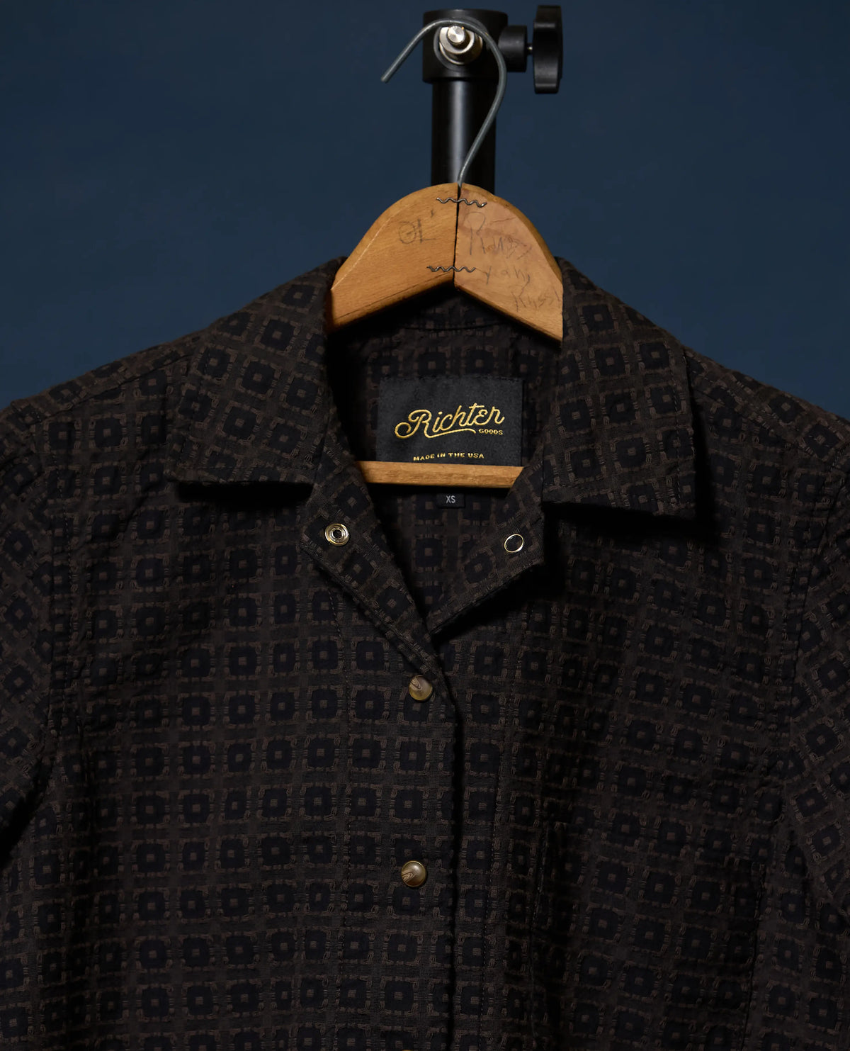A close-up of a dark patterned button-up shirt hanging on a wooden hanger against a dark blue background. The shirt has a small-scale geometric pattern in black and dark brown, with a visible 'Richter' neck label.
