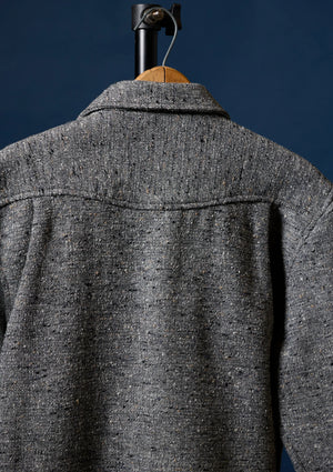 Close-up of the back of a textured gray tweed Wool shirt fior men hanging on a wooden hanger against a dark navy background.