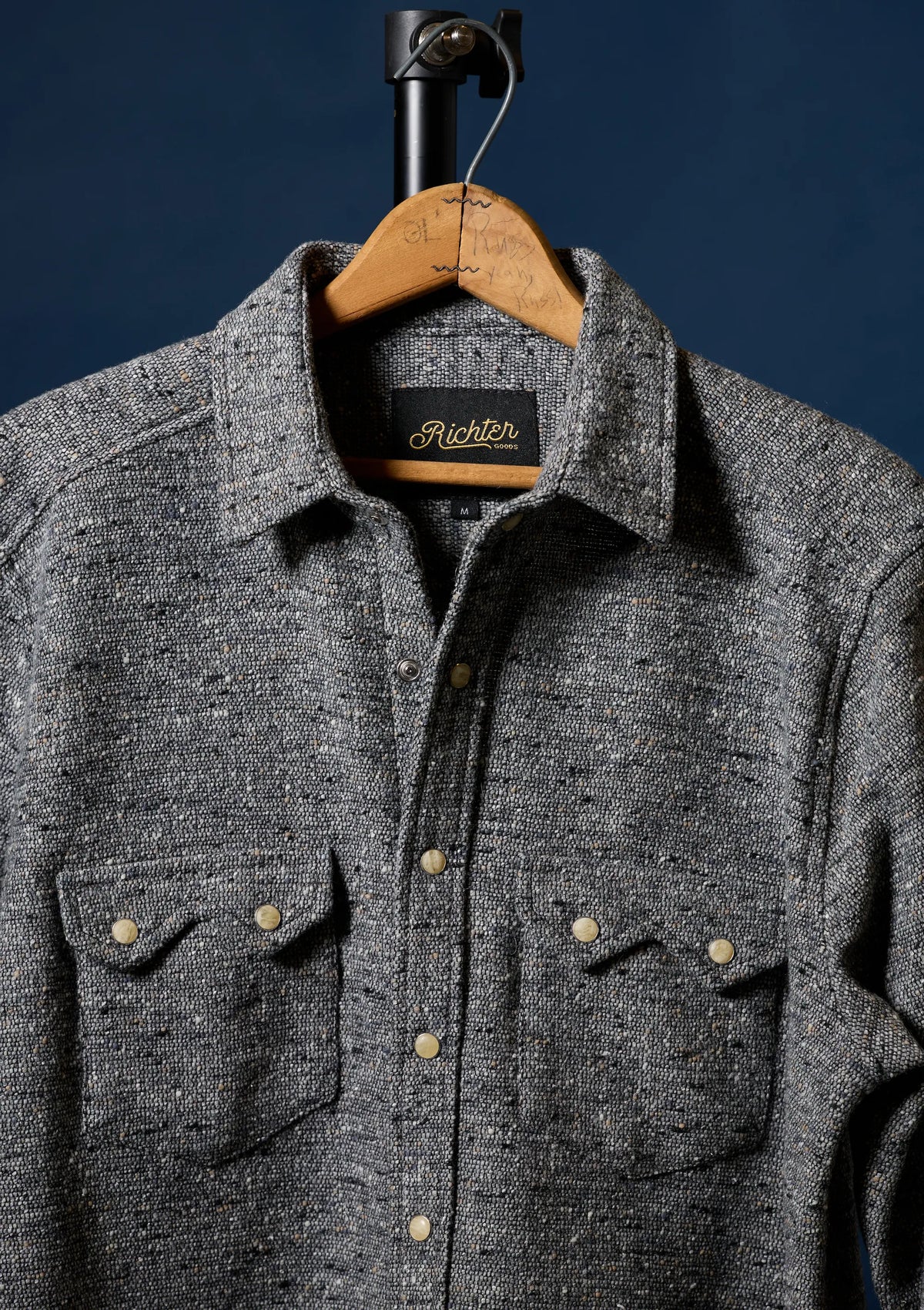A dark gray heathered wool button-up shirt for men with two chest pockets and brass buttons, hanging on a wooden hanger against a dark navy blue background. The brand label inside the collar reads 'Richter'.