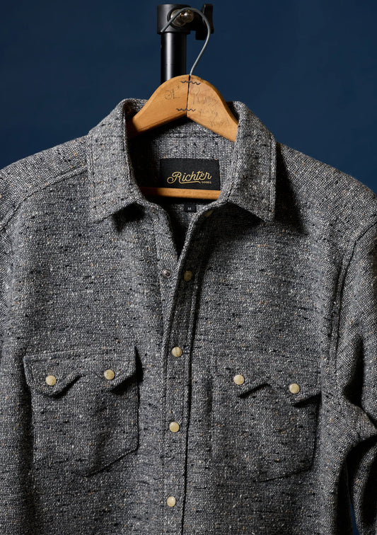 A dark gray heathered wool button-up shirt for men with two chest pockets and brass buttons, hanging on a wooden hanger against a dark navy blue background. The brand label inside the collar reads 'Richter'.