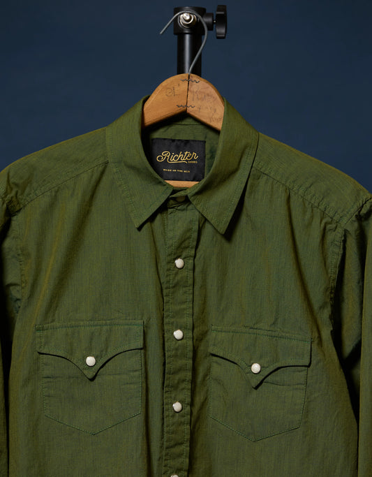 An olive green long-sleeve button-up shirt by Richter, hanging on a wooden hanger against a dark navy blue background.