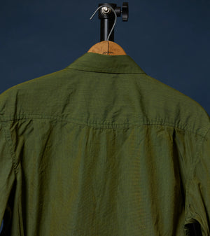 An olive green long-sleeve shirt shown from the back, hanging on a wooden hanger with a black stand against a dark navy background.