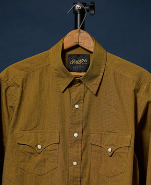 A close-up of a brown textured button-down shirt hanging on a wooden hanger against a dark navy background. The shirt features two buttoned chest pockets and a visible 'Richter Goods'
label.