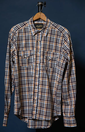 A long-sleeve plaid button-up shirt hanging on a wooden hanger against a dark navy background. The shirt features a brown, white, and black plaid pattern with two buttoned chest pockets.
