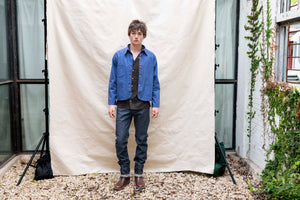 Person wearing a blue jacket and dark jeans standing in front of a white backdrop outdoors.