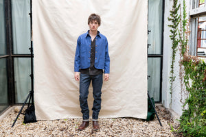 Person wearing a blue jacket and dark jeans standing in front of a white backdrop outdoors.
