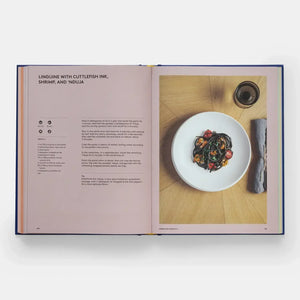 Open book showing a recipe for linguine with cuttlefish ink, shrimp, and nduja on a wooden surface.