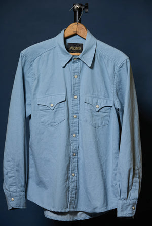 A light blue long-sleeve button-up shirt with two chest pockets, hanging on a wooden hanger against a dark navy background.