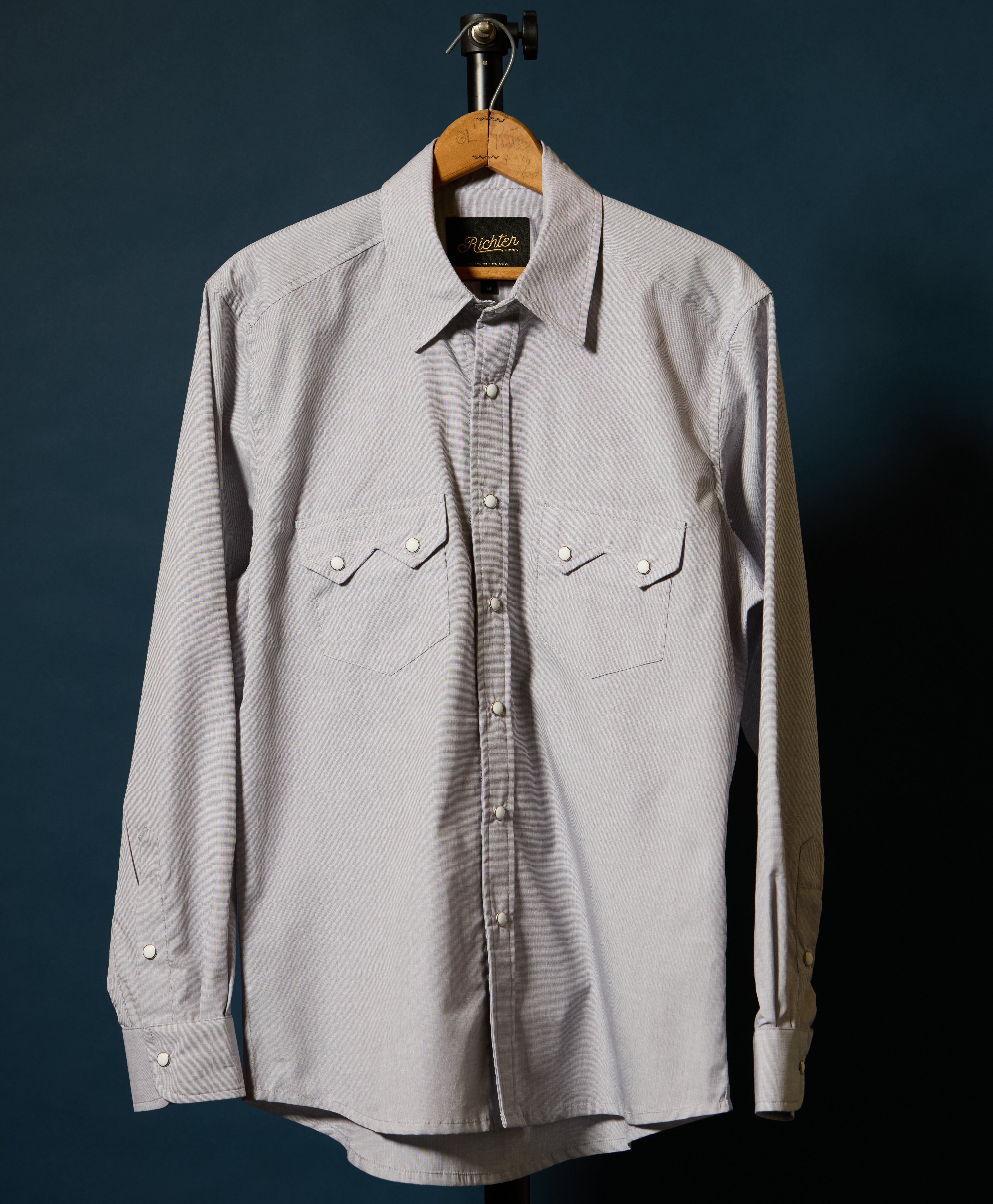 A Beige long-sleeve button-up shirt with two chest flap pockets, hanging on a wooden hanger against a dark blue background.