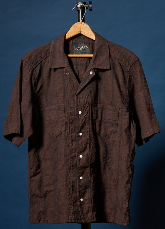 A dark brown short-sleeve button-up shirt with a textured crinkle fabric, hanging on a wooden hanger against a dark blue background.