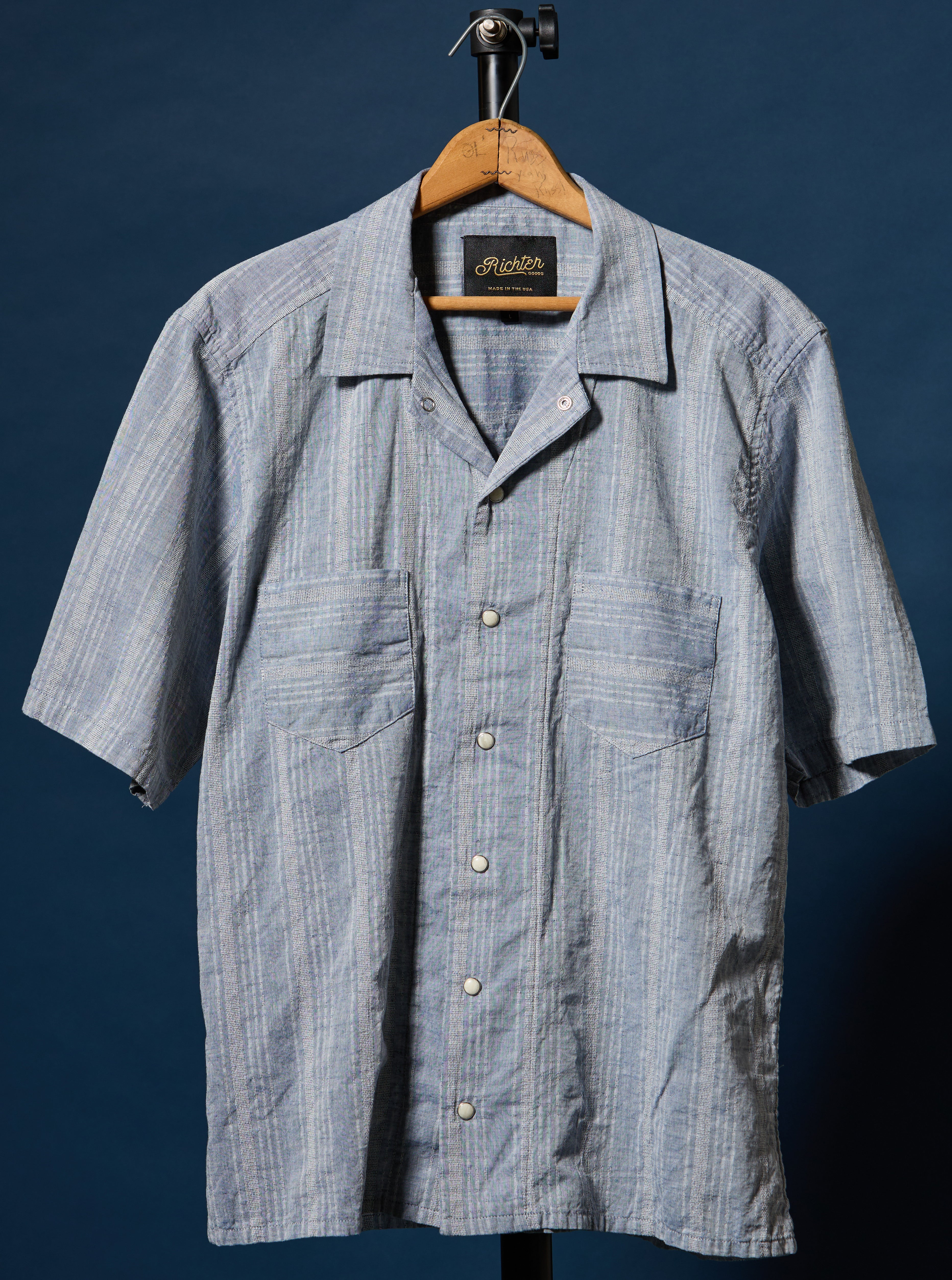 A short-sleeve button-up shirt with a light blue and white vertical stripe pattern, hanging on a wooden hanger against a dark blue background. The shirt features a camp collar, a button-down front, and two chest pockets.