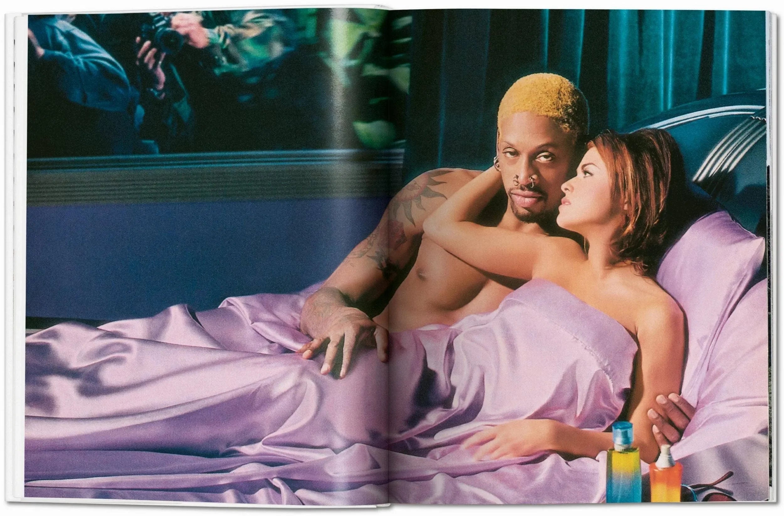 Two people with tattooed arms and piercings lying in bed under purple satin sheets, with colorful bottles on bedside table; bedroom scene