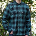 A model wearing a dark green and black plaid long-sleeve button-up shirt, dark blue jeans,  standing outdoors against a blurred background of green foliage.