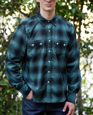 A model wearing a dark green and black plaid long-sleeve button-up shirt, dark blue jeans,  standing outdoors against a blurred background of green foliage.