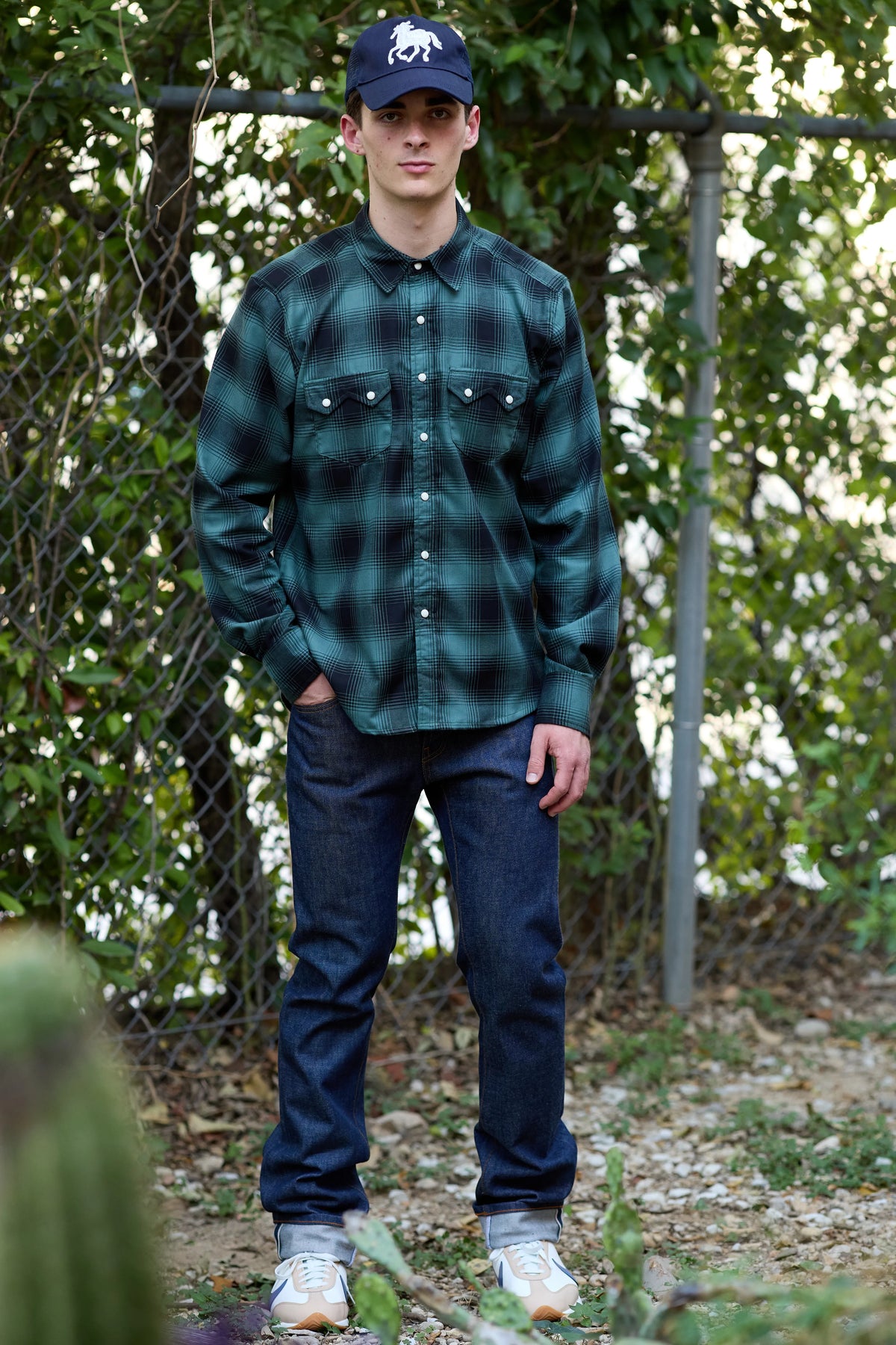 A model wearing a Richter Goods' dark green and black plaid button-down shirt, dark blue jeans, a navy baseball cap, and white sneakers, standing outdoors in front of a chain-link fence and trees.