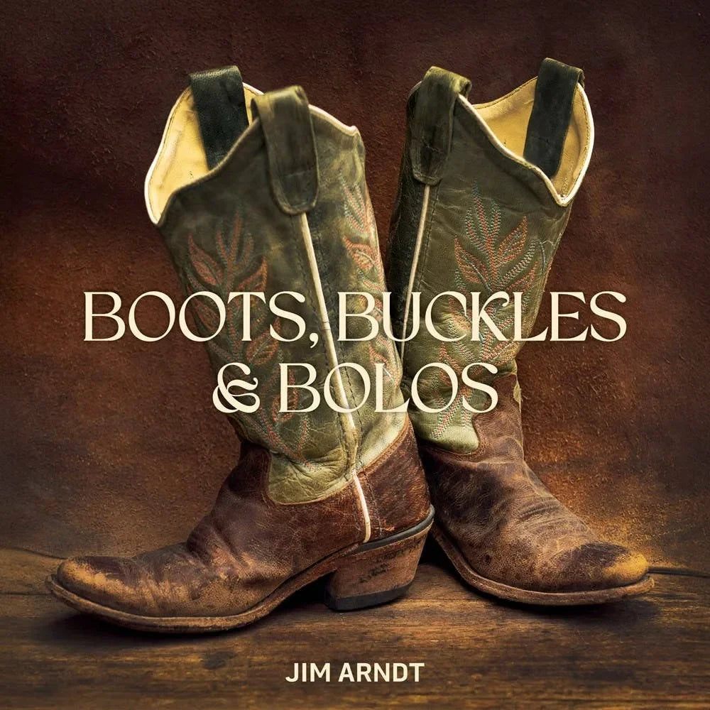Pair of vintage cowboy boots with decorative patterns on a wooden surface, featuring the text 'Boots, Buckles & Bolos' and 'Jim Arndt'.
