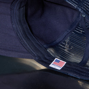 Close-up of a dark navy trucker hat showing the mesh back, stitched fabric edge, and a small white 'MADE IN USA' patch with an American flag.