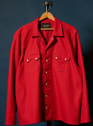 A bright red long-sleeve button-up shirt with two sawtooth chest pockets and brass-tone buttons, hanging on a wooden hanger against a dark blue background.