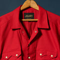 A bright red button-up shirt jacket by Richter Goods, hanging on a wooden hanger against a dark blue background. The jacket features a classic collar, front button closure, and two buttoned sawtooth pockets.