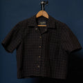 A short-sleeve button-up shirt with a dark brown and black geometric pattern, hanging on a wooden hanger against a dark blue background.