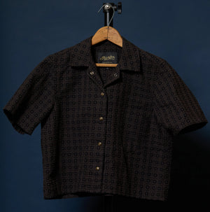 A short-sleeve button-up shirt with a dark brown and black geometric pattern, hanging on a wooden hanger against a dark blue background.