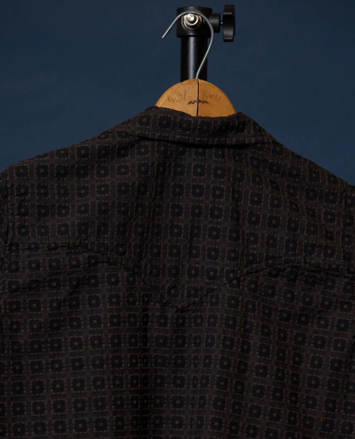 A dark patterned shirt displayed on a wooden hanger against a dark blue background.
