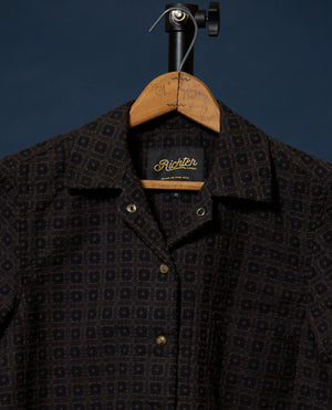 A close-up of a dark patterned button-up shirt hanging on a wooden hanger against a dark blue background. The shirt has a small-scale geometric pattern in black and dark brown, with a visible 'Richter' neck label.