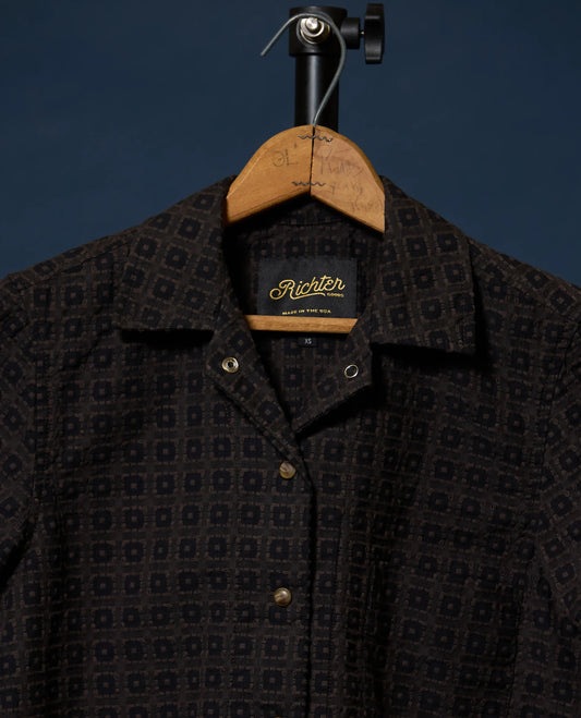 A close-up of a dark patterned button-up shirt hanging on a wooden hanger against a dark blue background. The shirt has a small-scale geometric pattern in black and dark brown, with a visible 'Richter' neck label.