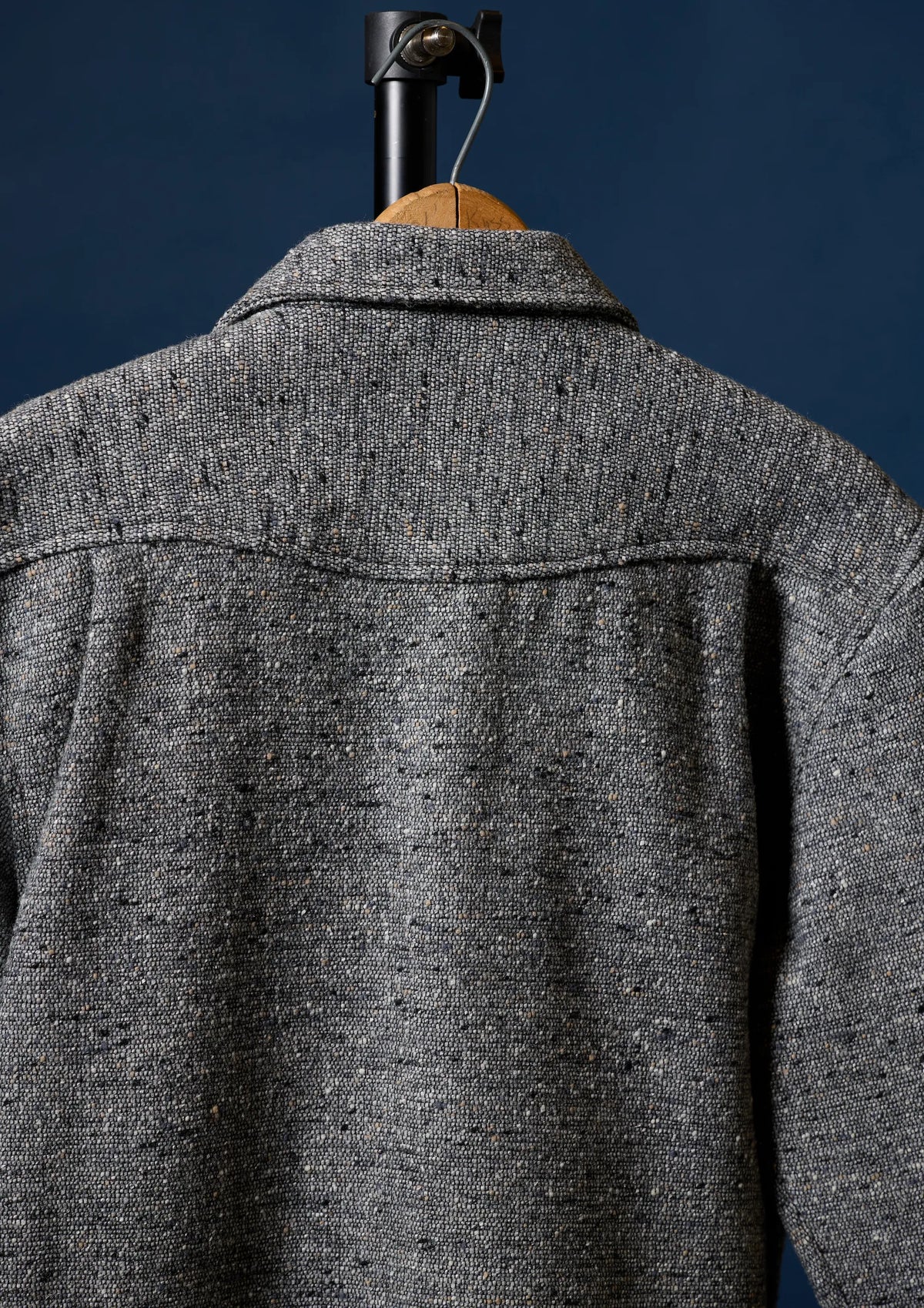 Close-up of the back of a textured gray tweed Wool shirt fior men hanging on a wooden hanger against a dark navy background.
