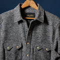 A dark gray heathered wool button-up shirt for men with two chest pockets and brass buttons, hanging on a wooden hanger against a dark navy blue background. The brand label inside the collar reads 'Richter'.