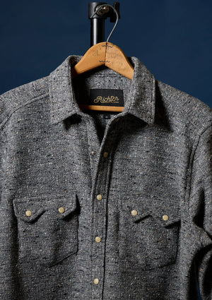 A dark gray heathered wool button-up shirt for men with two chest pockets and brass buttons, hanging on a wooden hanger against a dark navy blue background. The brand label inside the collar reads 'Richter'.