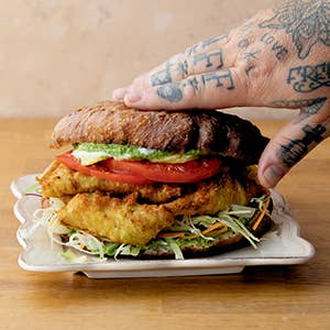 Hand with tattoos holding a large sandwich on a wooden table