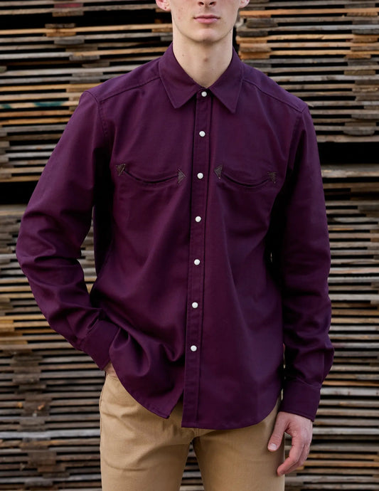 A man wearing a long-sleeve button-up shirt in Maroon / Burgundy, paired with tan pants, standing in front of a rustic wooden slat wall.
