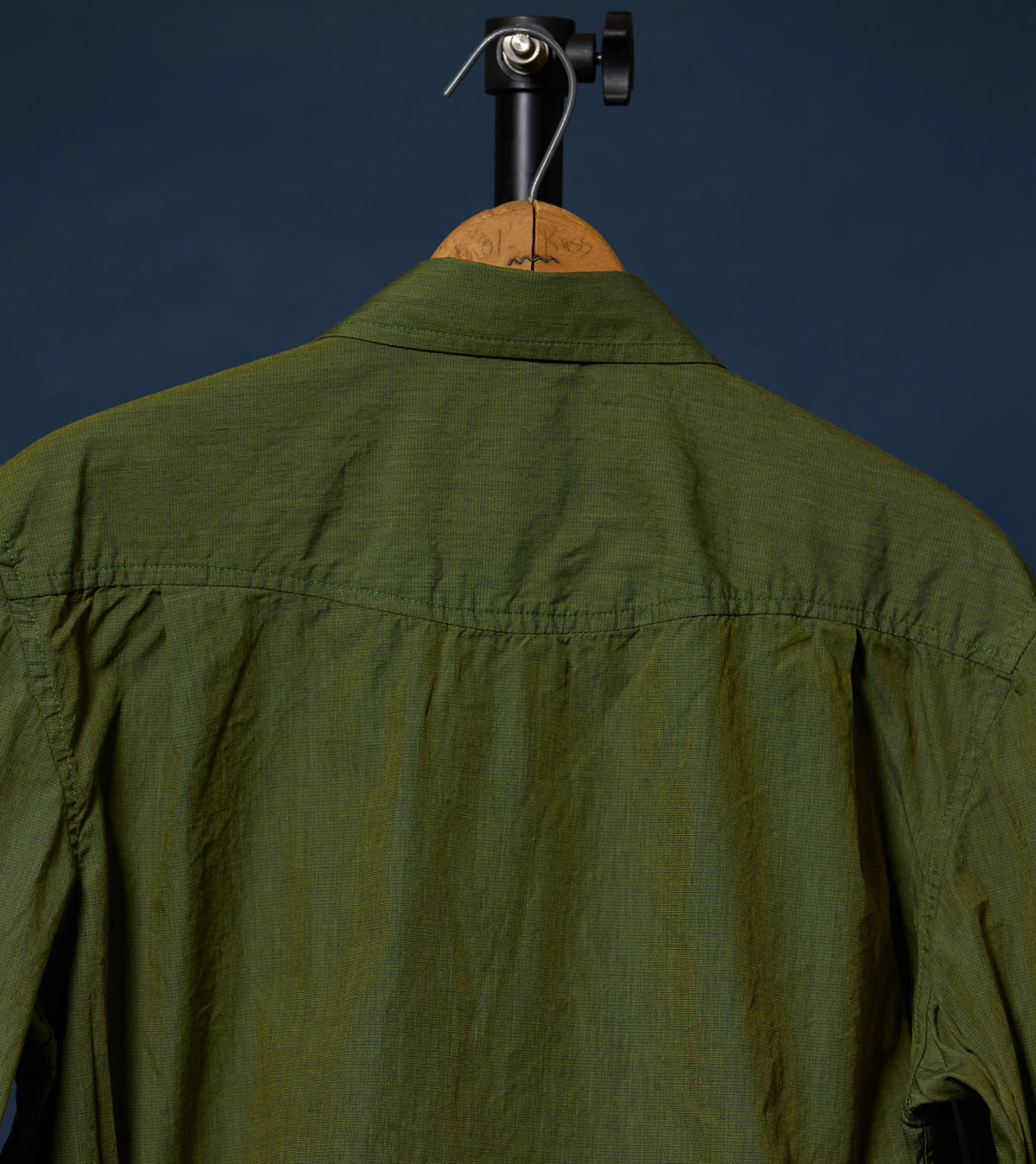 An olive green long-sleeve shirt shown from the back, hanging on a wooden hanger with a black stand against a dark navy background.