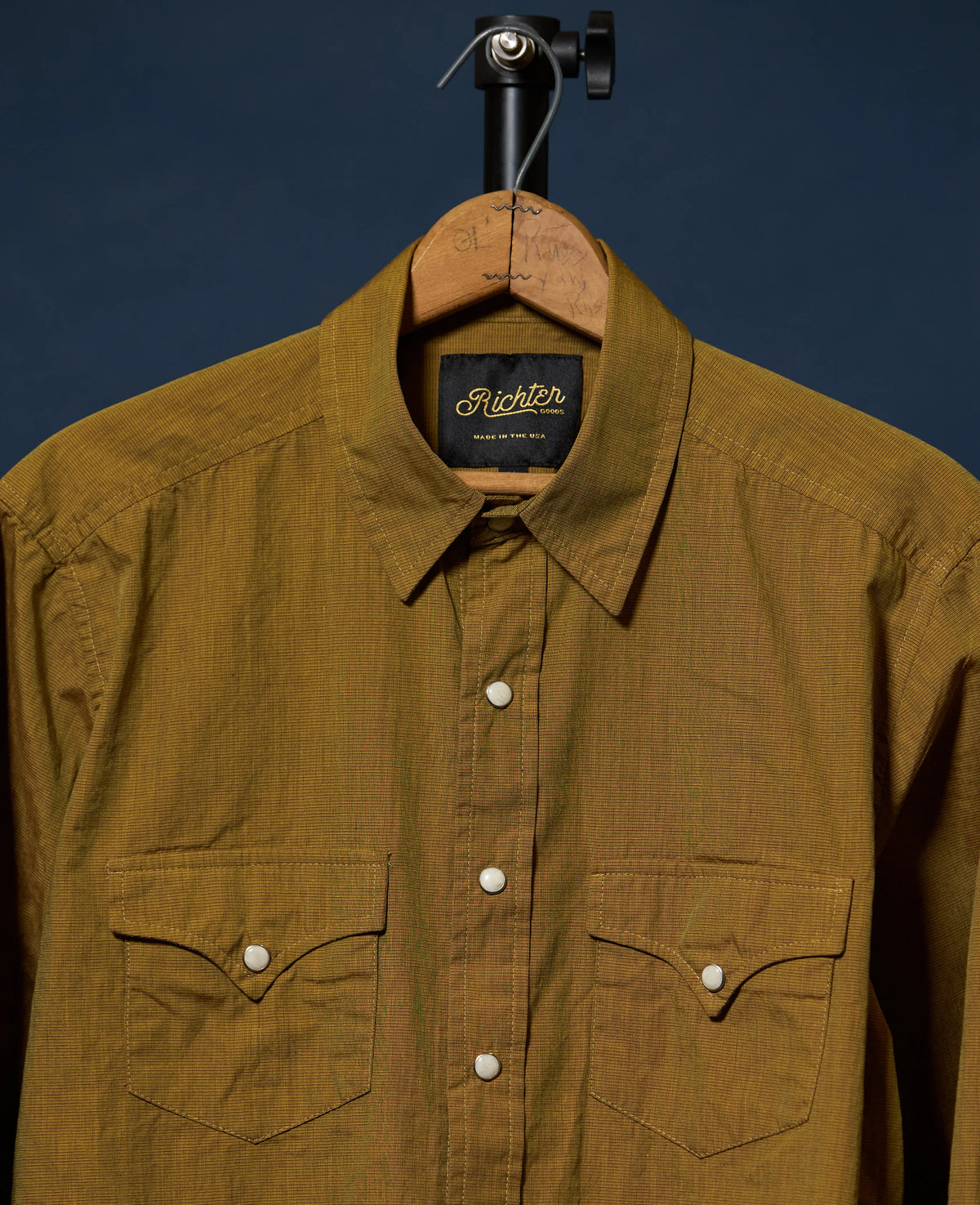 A close-up of a brown textured button-down shirt hanging on a wooden hanger against a dark navy background. The shirt features two buttoned chest pockets and a visible 'Richter Goods' 
label.
