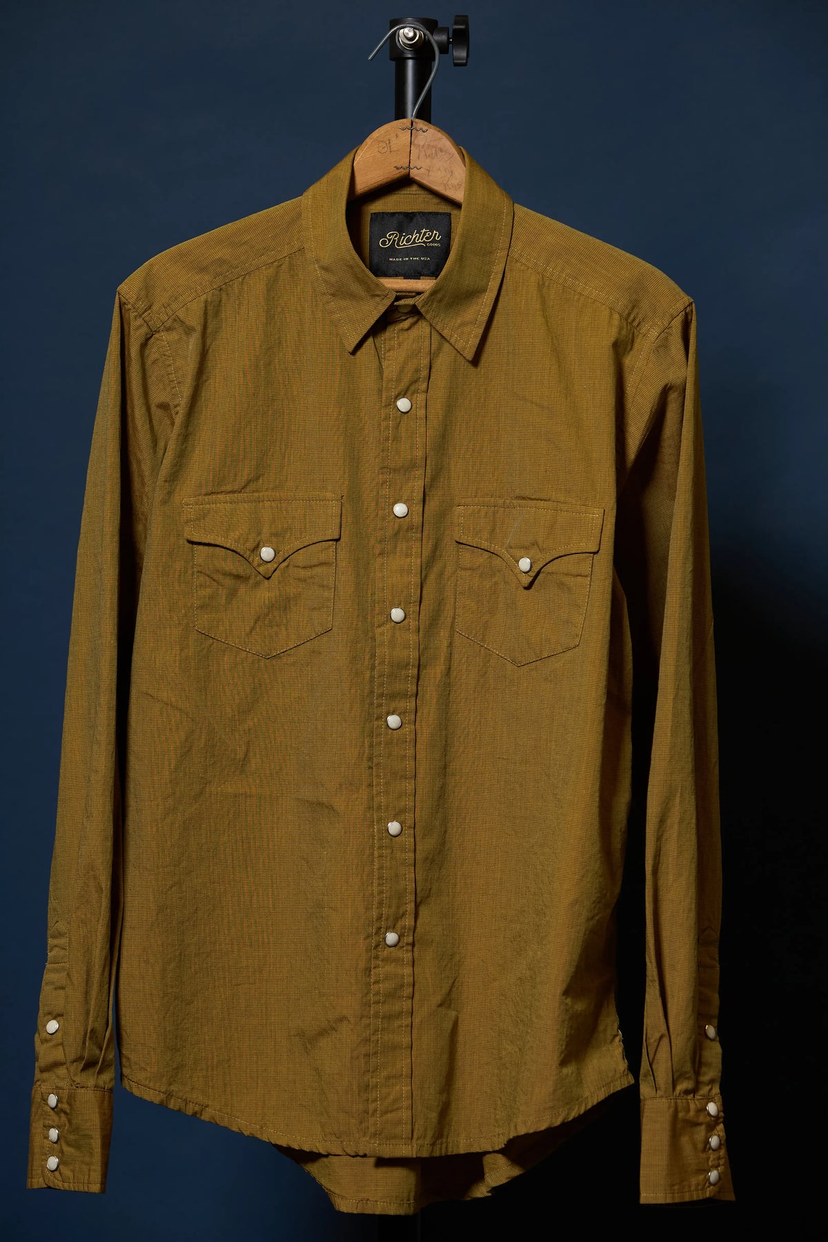 A long-sleeve button-up shirt in a textured mustard yellow fabric, hanging on a wooden hanger against a dark navy blue background.
