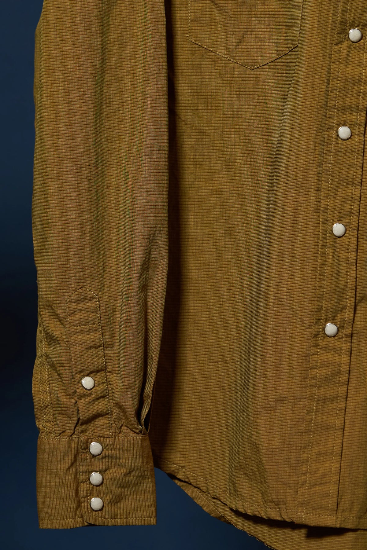 Close-up of a mustard yellow linen shirt with white buttons, showing the chest pocket, button-down front, and a buttoned cuff against a dark blue background.