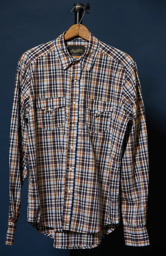 A long-sleeve plaid button-up shirt hanging on a wooden hanger against a dark navy background. The shirt features a brown, white, and black plaid pattern with two buttoned chest pockets.