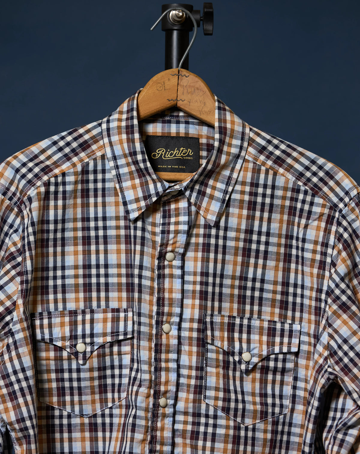 A plaid button-up shirt by Richter Goods hanging on a wooden hanger against a dark navy background. The shirt features a checkered pattern of black, brown, and white, with two buttoned chest pockets.