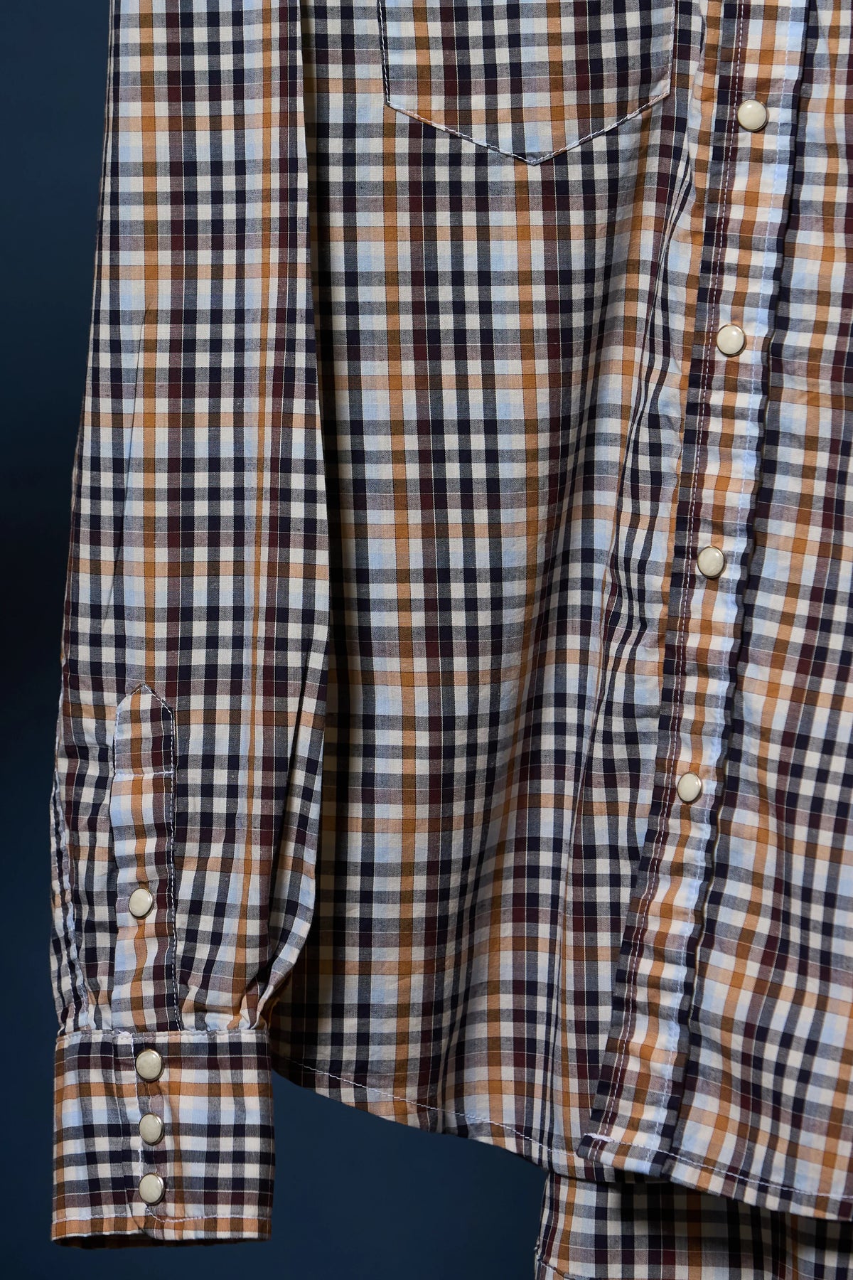 A close-up of a long-sleeve plaid button-up shirt with a chest pocket, featuring a checkered pattern of camel, brown, white, and black, with light-colored buttons.