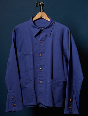 Barista Chore Coat in Washed Herringbone with brown pearl snaps on a hanger against a dark background