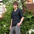 Model wearing a black shirt and gray pants standing against a green ivy-covered wall.