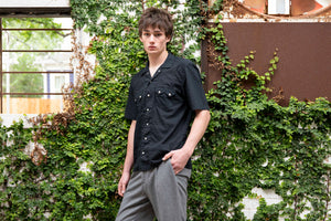 Model wearing a black shirt and gray pants standing against a green ivy-covered wall.