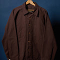 A dark brown utility overshirt jacket with a button-up front, collar, and chest pocket, hanging on a wooden hanger against a dark blue background.