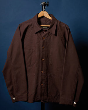 A dark brown utility overshirt jacket with a button-up front, collar, and chest pocket, hanging on a wooden hanger against a dark blue background.