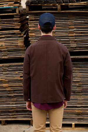 A man viewed from behind wearing a dark brown utility jacket, a navy blue baseball cap, a purple shirt, and khaki pants, standing in front of a stack of wooden pallets.